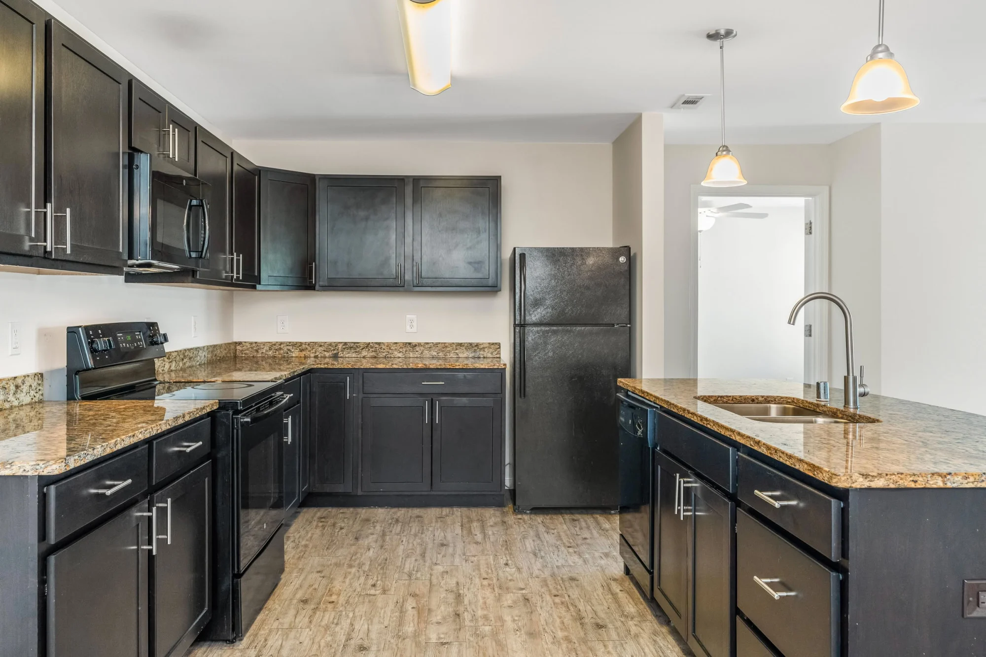 Roomy apartment kitchen with dark espresso cabinets, granite countertops, black appliances, a center island with stainless sink, pendant lighting, and light wood-style flooring.