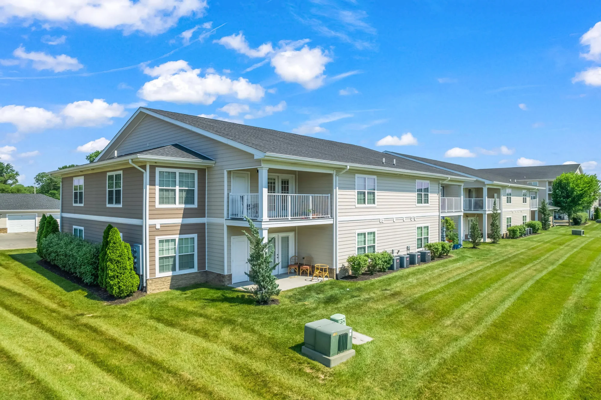 Two-story tan apartment building with covered porches and balconies, lush green lawn, neatly trimmed shrubs, and bright blue sky with scattered white clouds overhead.