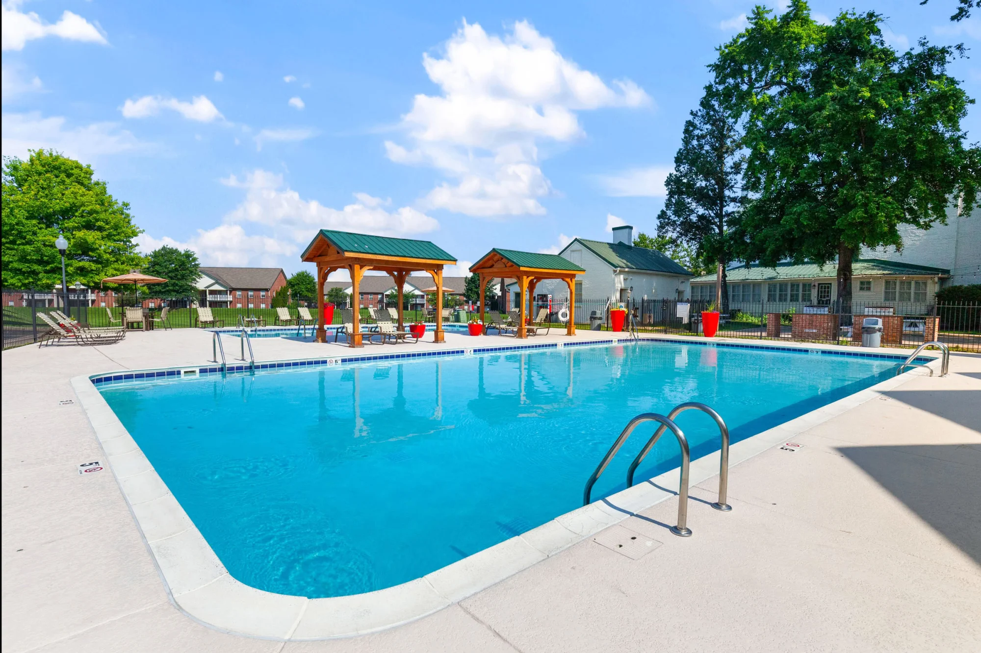 Sparkling blue resort-style swimming pool with two wooden pergolas, lounge chairs, and bright red planters set against a backdrop of trees and apartment buildings under a blue sky.